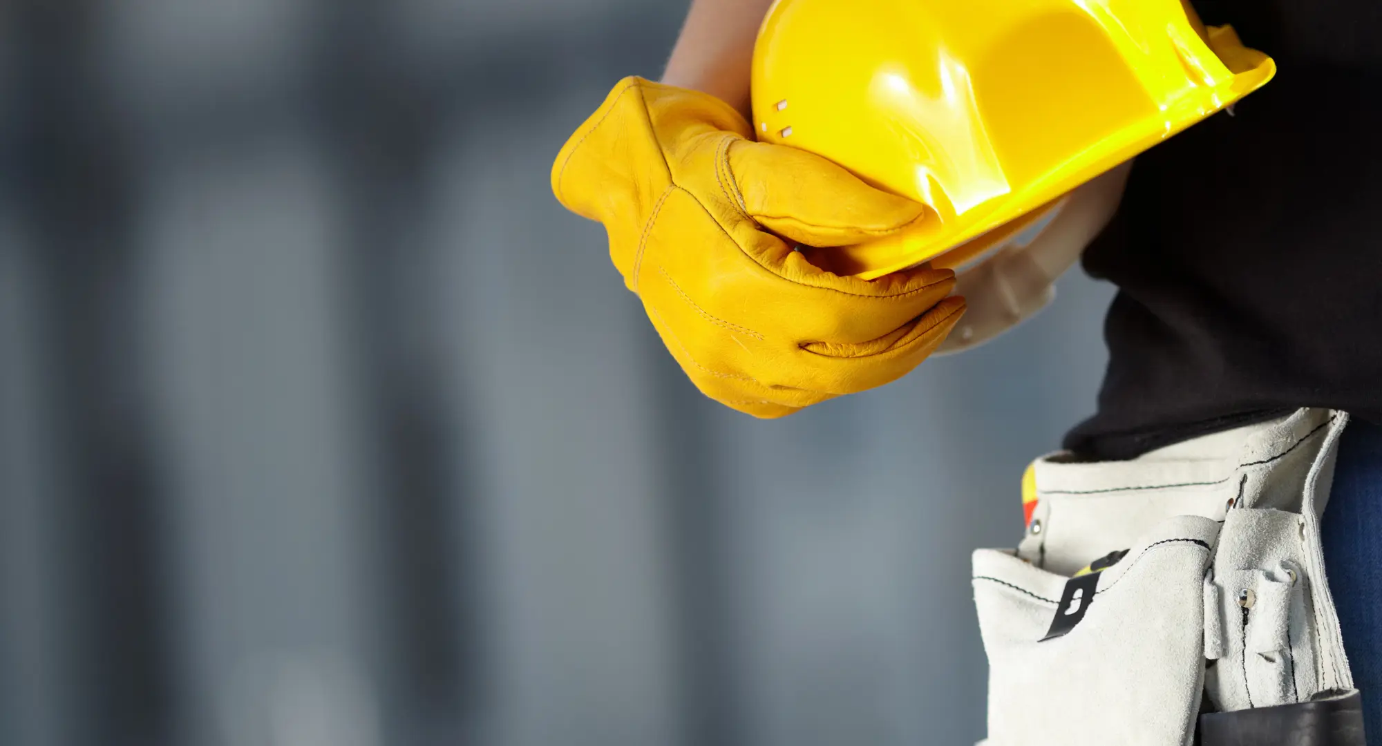 Construction worker wearing yellow gloves and holding a yellow hard hat, with a tool belt visible at the waist.