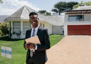 Real estate agent in suit holding clipboard in front of house with 'For Sale' sign and scenic backdrop