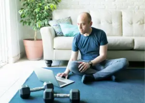 Man sitting cross-legged on the floor at home using a laptop, with dumbbells nearby in front of a couch, representing online fitness or health activities.