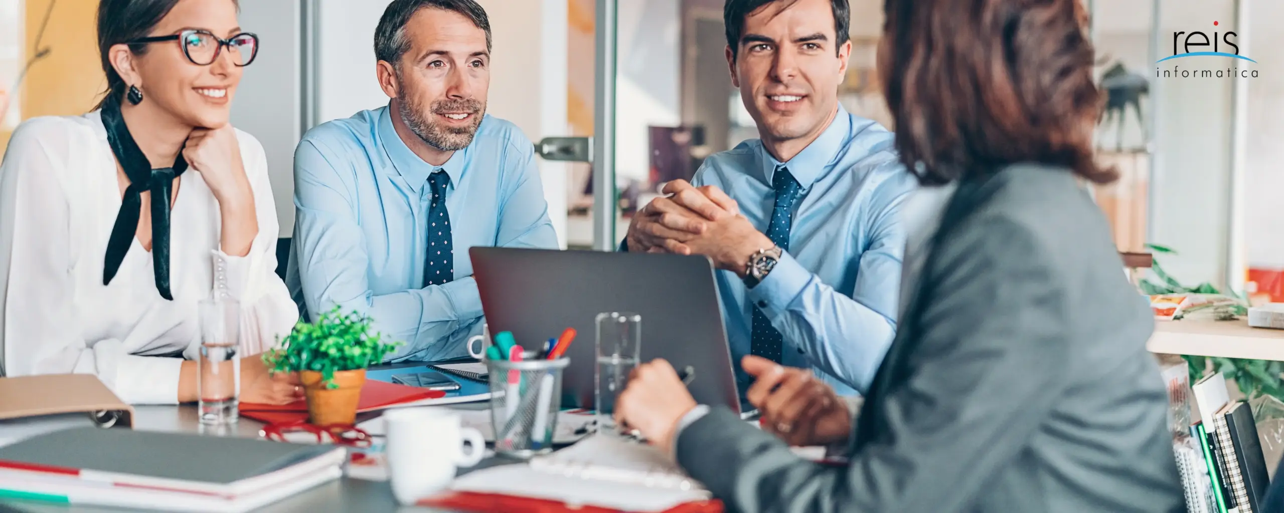 Business team in modern office discussing strategy around table with laptops and documents – Reis Informatica