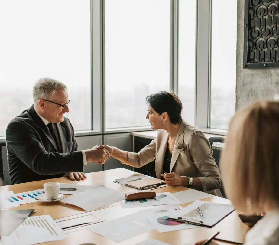 Business professionals shaking hands at a meeting table in a modern office with documents and charts spread out.